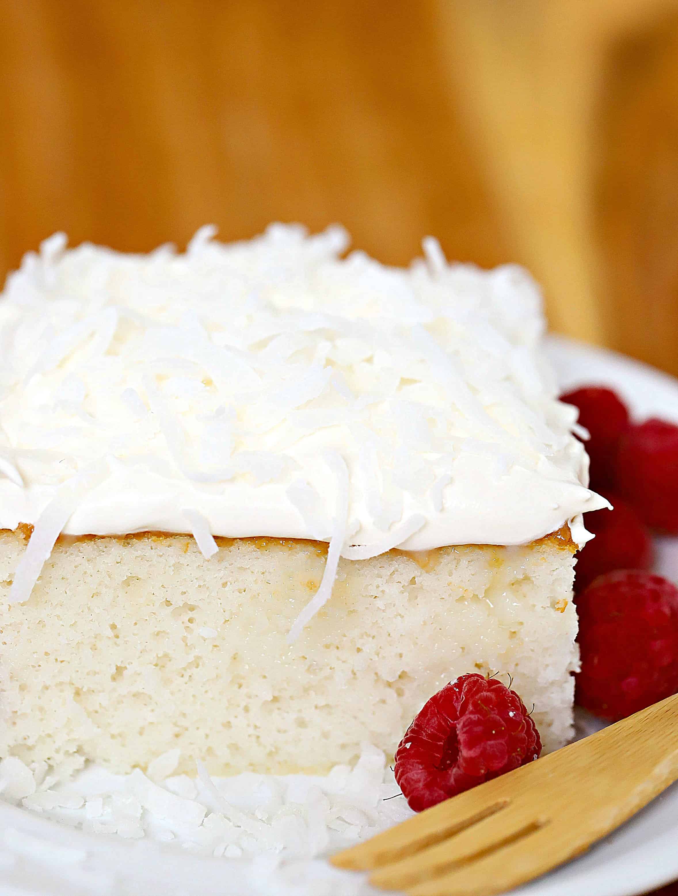 Close up of a square slice of cake topped with whipped cream and coconut and a side of raspberries.