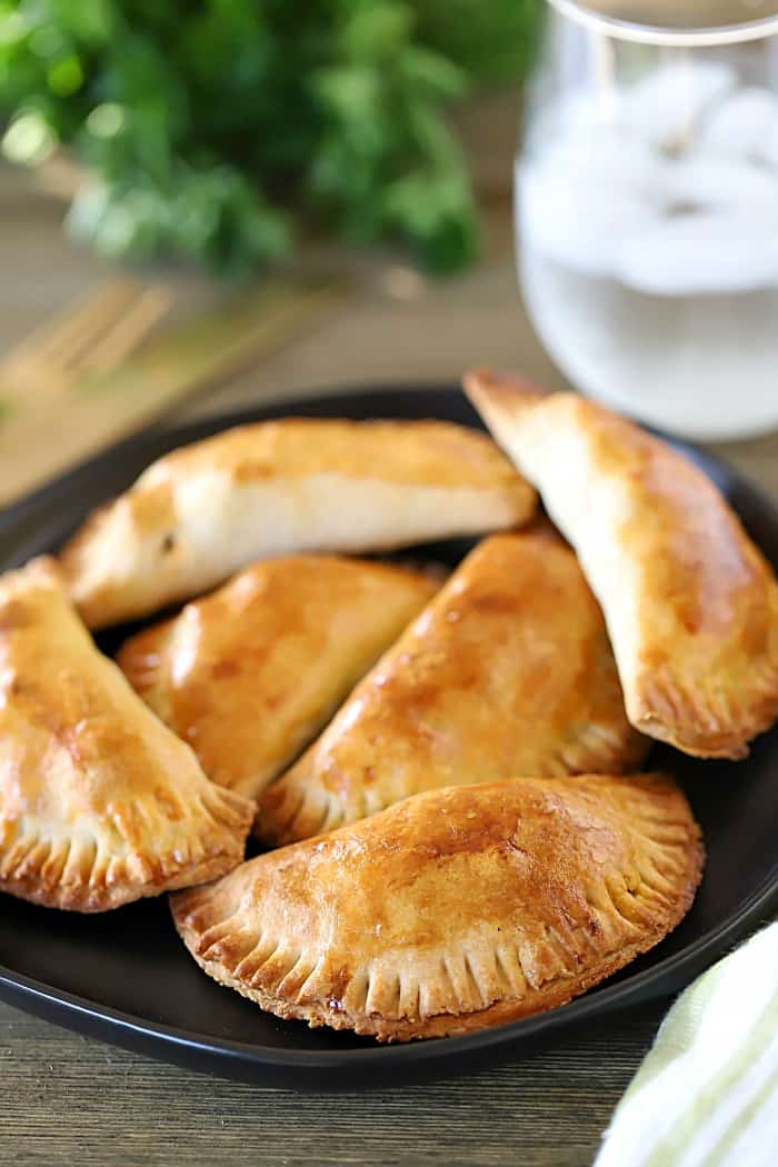 One black plate of four beef empanadas with a glass of water on the side.