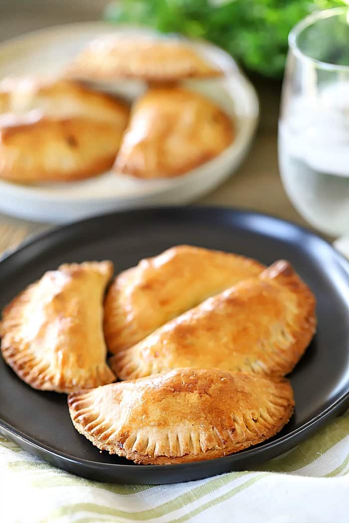 One black plate of four beef empanadas and one white plate in the background with a glass of water on the side.
